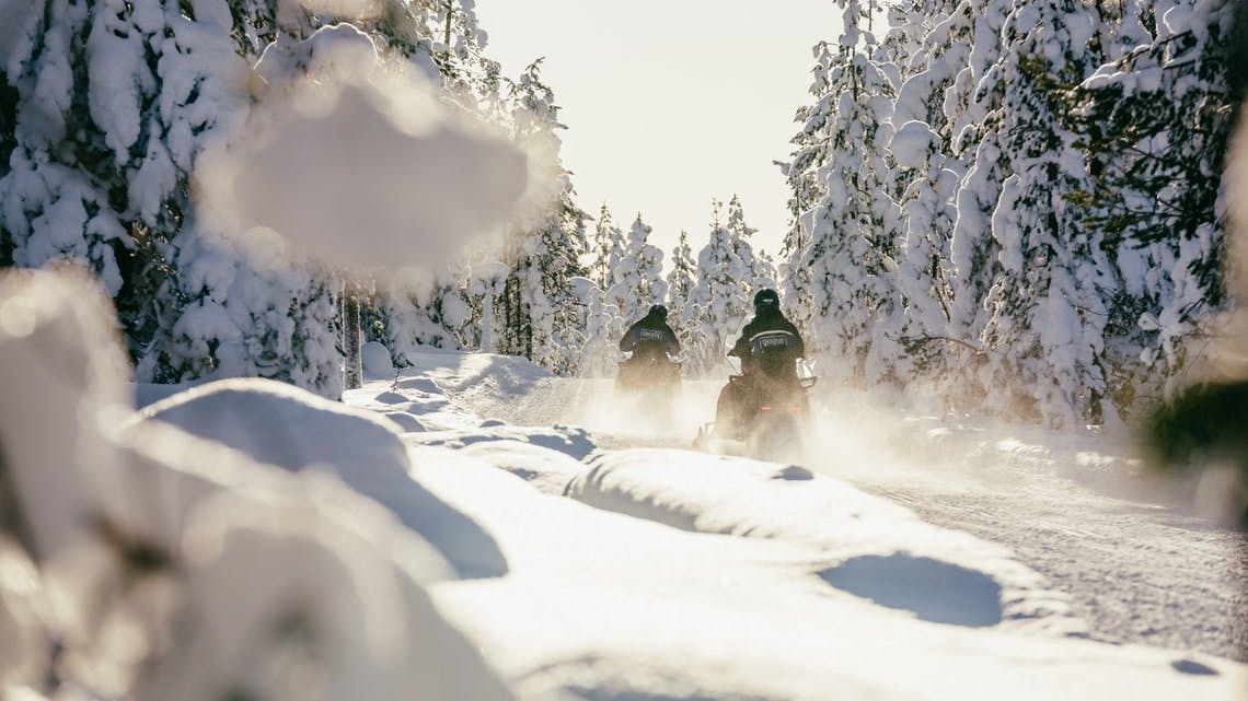 Δύο άνθρωποι οδηγούν οχήματα χιονιού σε ένα χιονισμένο μονοπάτι που περιβάλλεται από χιονισμένα δέντρα μια ηλιόλουστη μέρα.