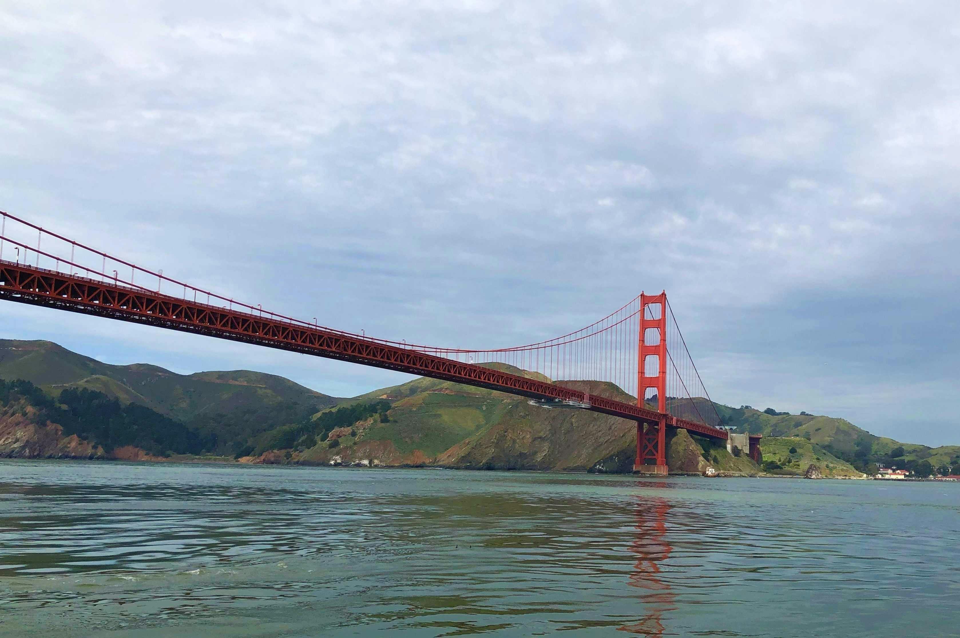 The Golden Gate Bridge stretches over calm waters with green hills in the background under a cloudy sky.