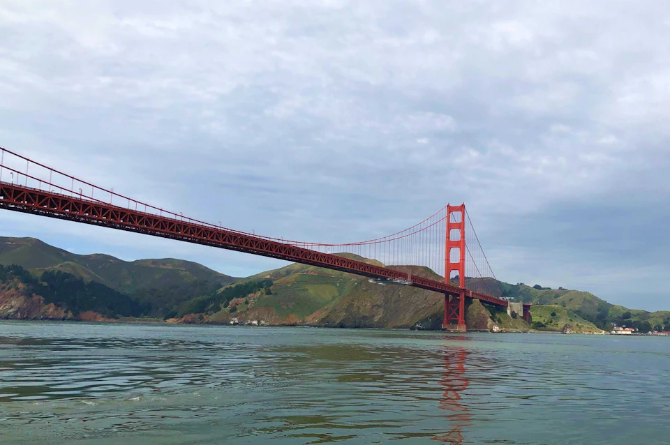 The Golden Gate Bridge stretches over calm waters with green hills in the background under a cloudy sky.