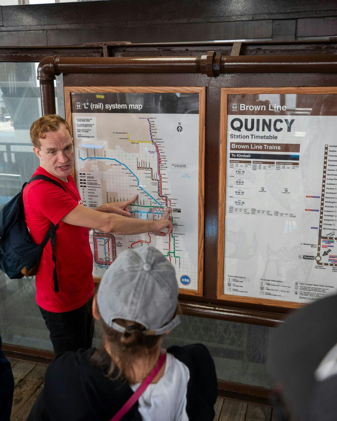 一名身穿红色衬衫的男子在与另一个人交谈时指着芝加哥“L”列车地图。 您可以看到昆西站的时间表。