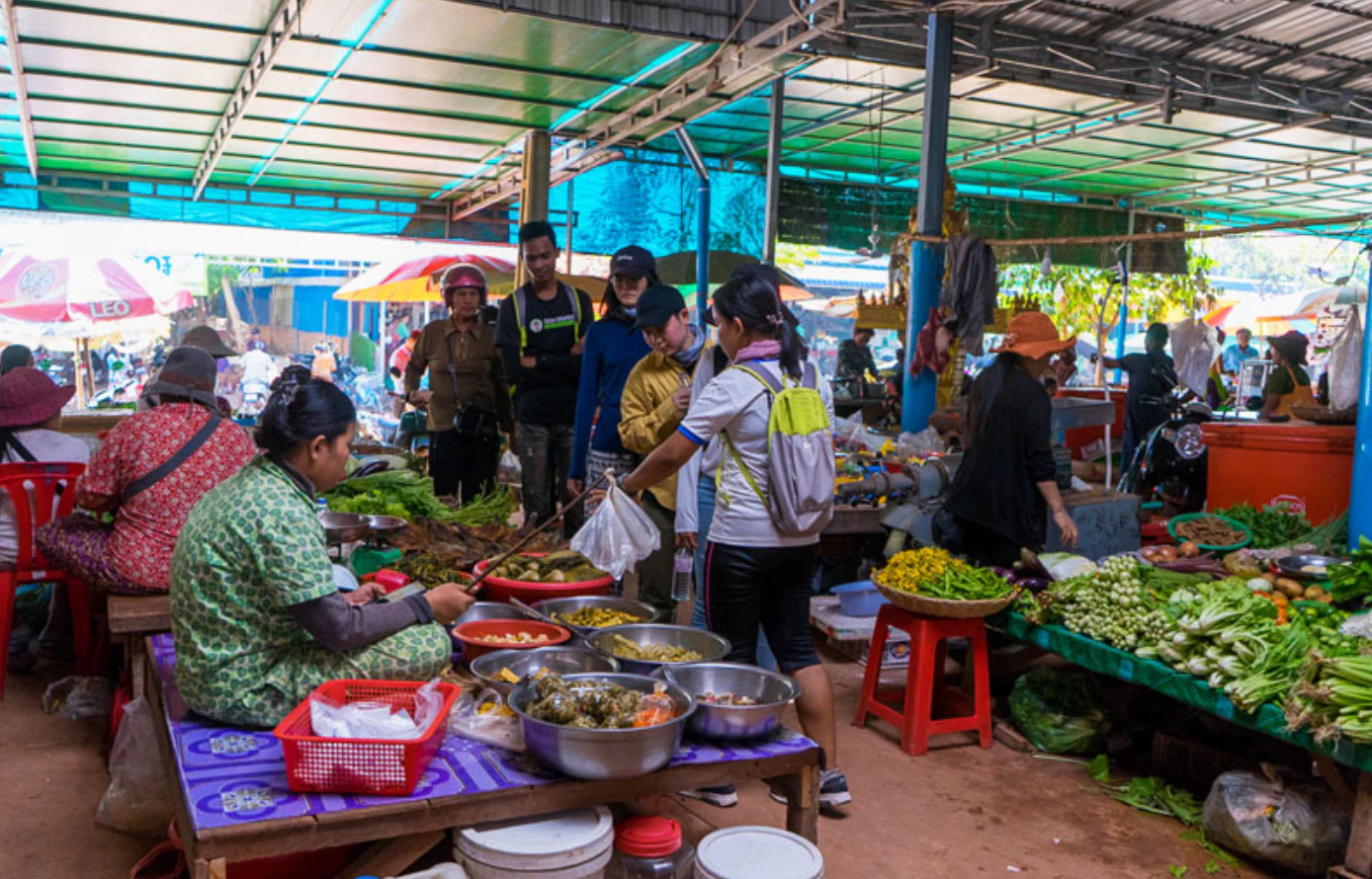 People interacting at a bustling market stall with various foods and goods under a metal canopy.