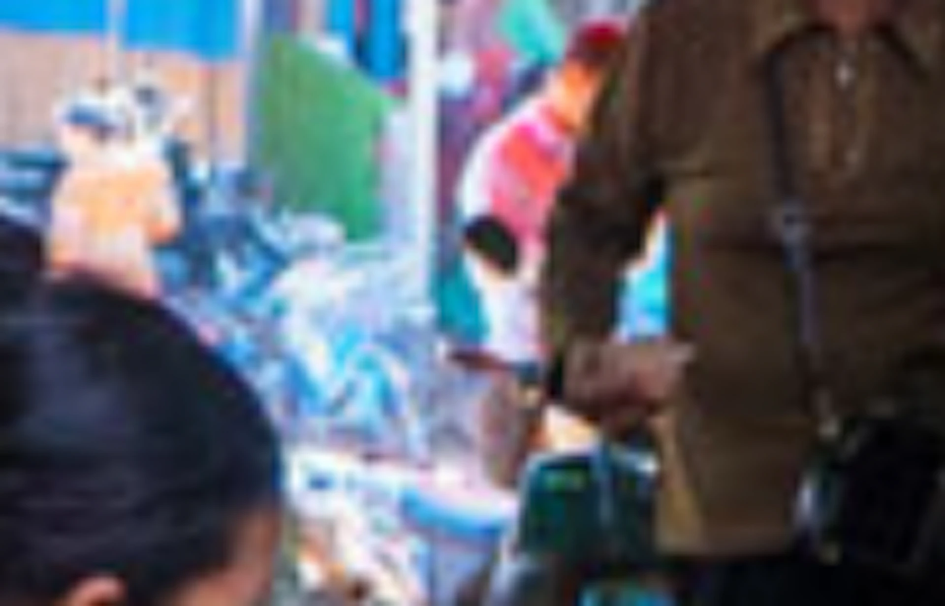 People interacting at a bustling market stall with various foods and goods under a metal canopy.