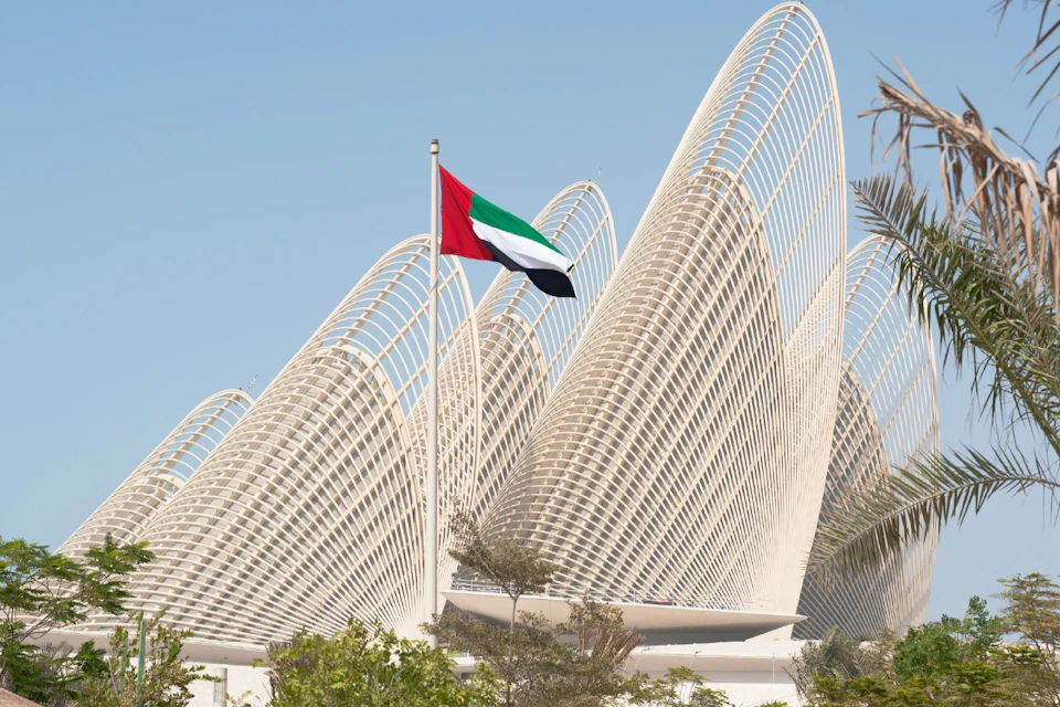 A United Arab Emirates flag on a pole with modern, curved architectural structures in the background under a clear sky.