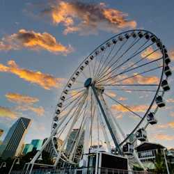 Ferris wheel against a sunset sky, with city buildings in the background and some clouds illuminated by the setting sun.