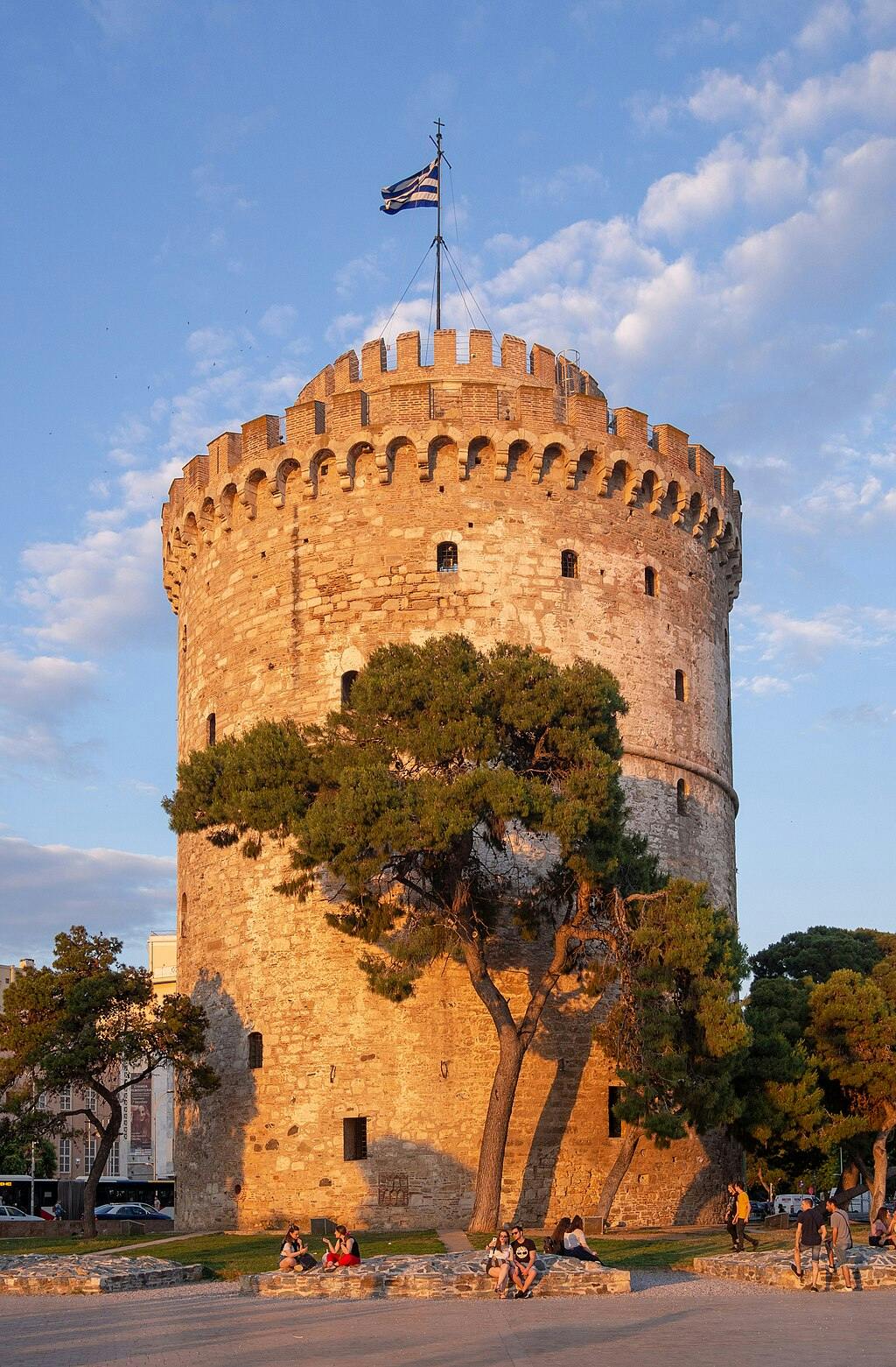 Una histórica torre de piedra con gente sentada y paseando alrededor de su base, árboles en primer plano y un cielo despejado por encima.