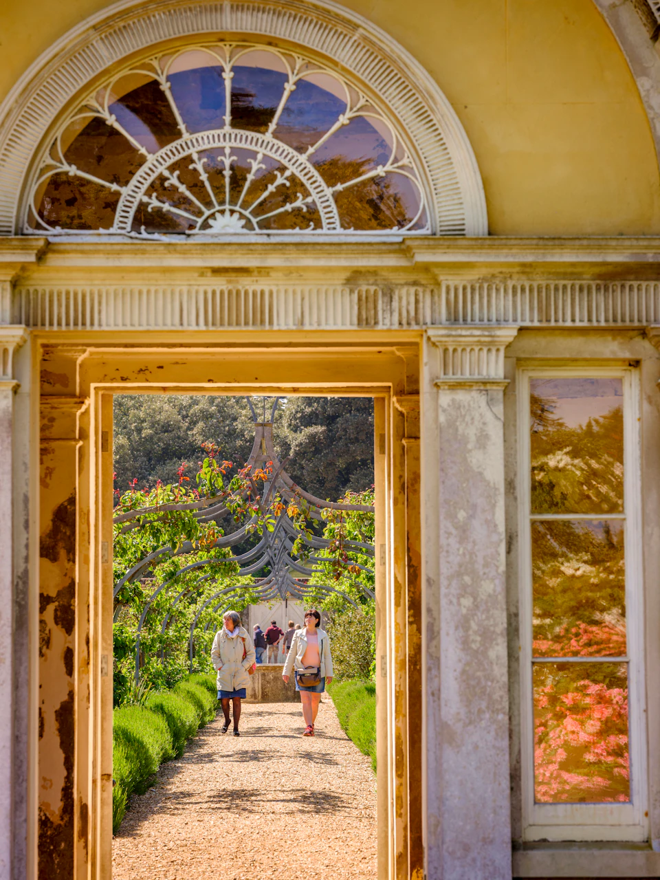 Two people walking through a garden archway, viewed through an ornate stone doorway with a glass window above. Trees and greenery surround them.
