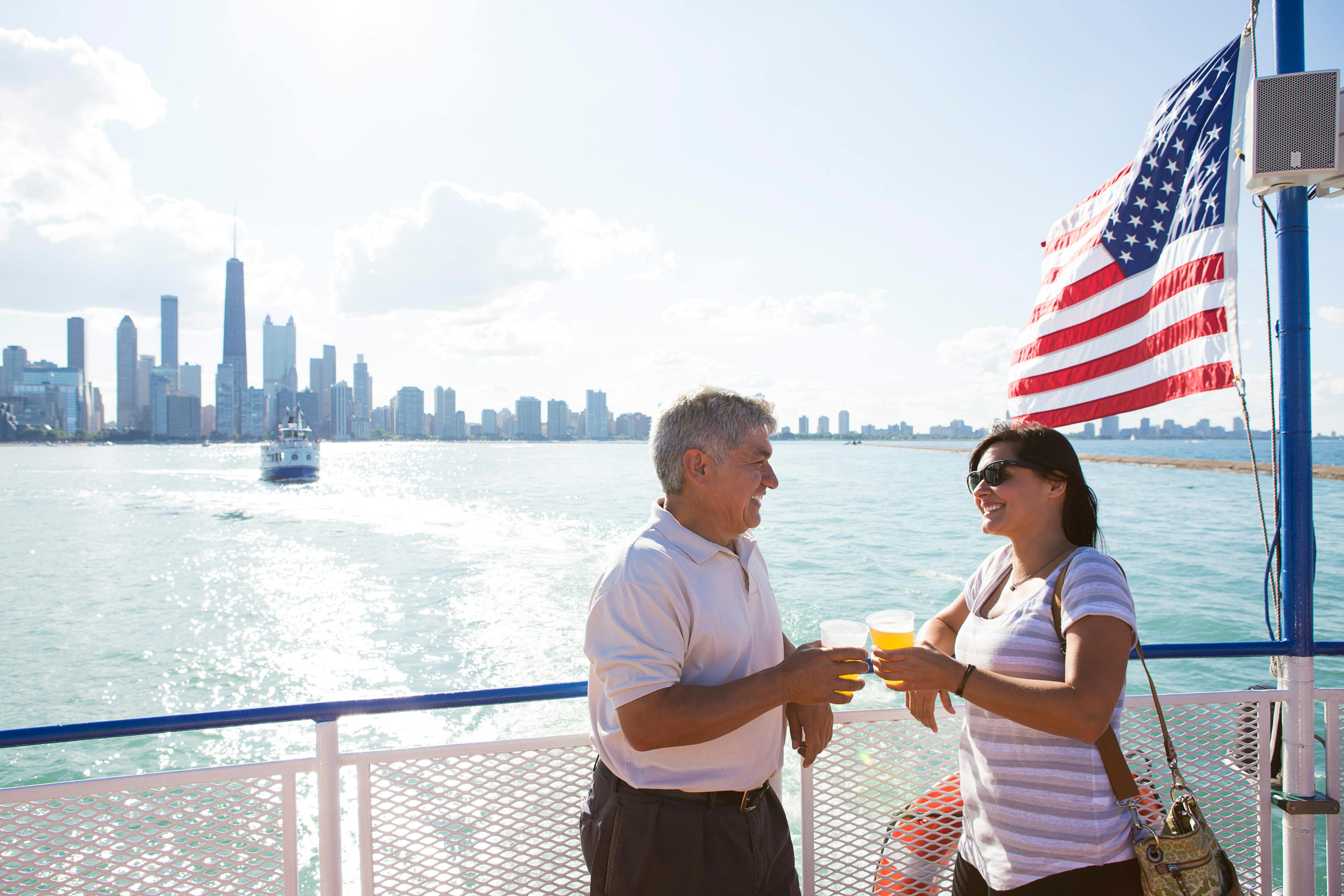 Dva lidé drží nápoje a usmívají se na lodi s panoramatem města, americkou vlajkou a vodou v pozadí.