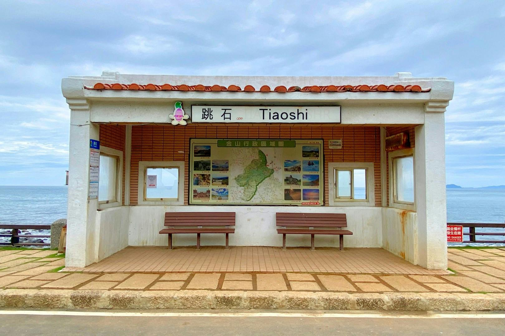 Małe nadmorskie schronisko z napisem „Tiaoshi”, z ławkami i mapą na ścianie, z widokiem na ocean pod zachmurzonym niebem.