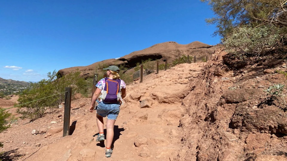 A person hiking up a rocky trail on a sunny day, wearing a backpack and casual clothes, surrounded by desert scenery.