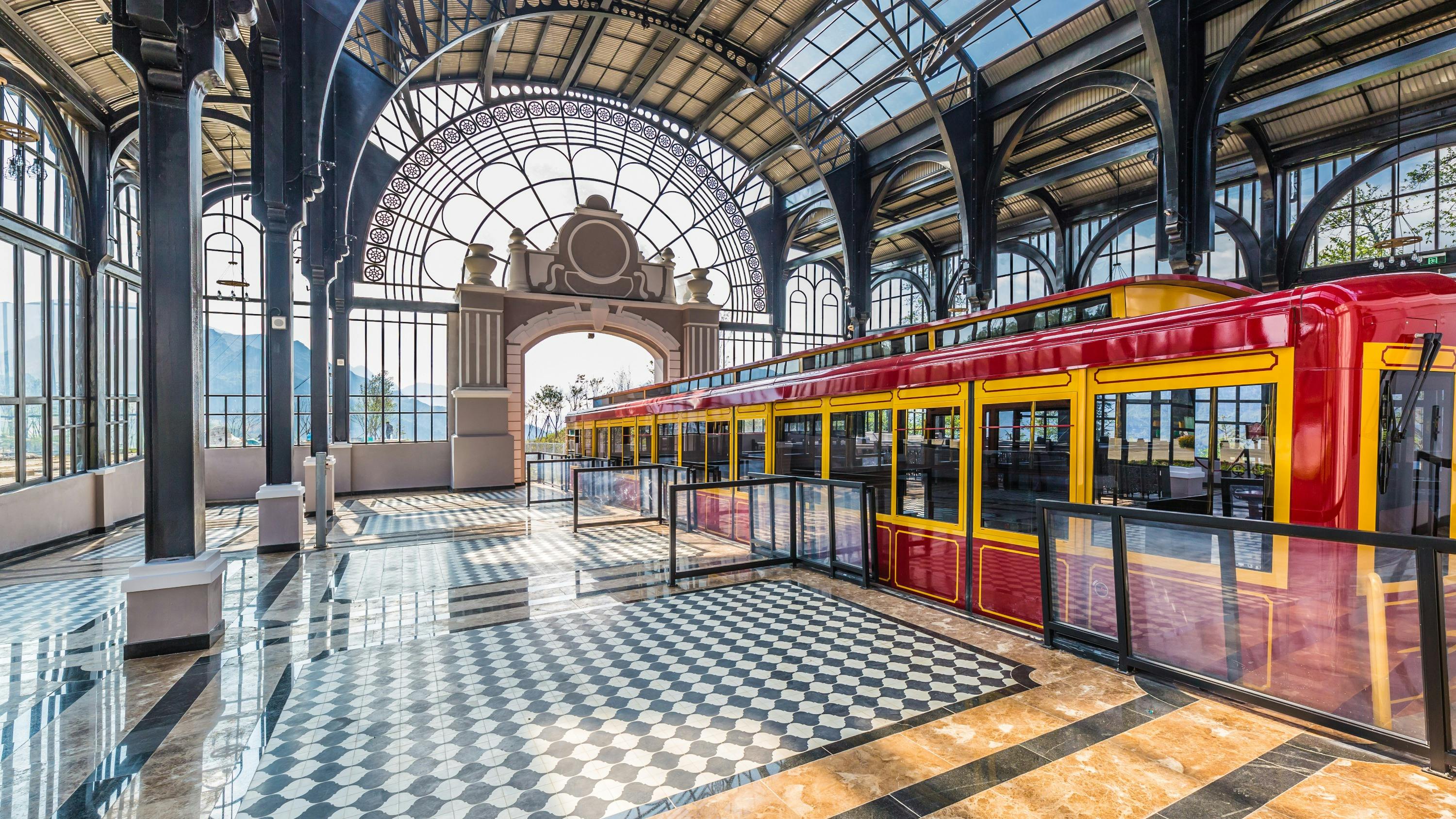 Colorful train inside a bright, ornate station with large windows and a checkered floor.