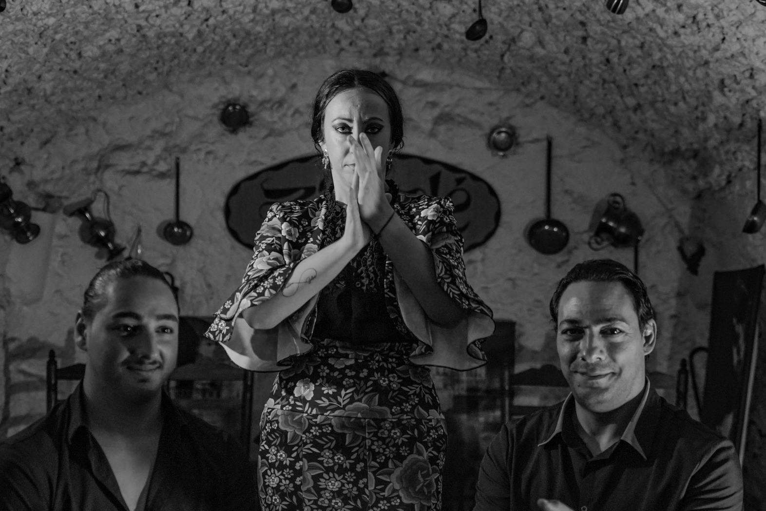 Ein Schwarz-Weiß-Bild von drei Personen, mit einer klatschenden Frau in der Mitte, in einem rustikalen Interieur mit hängenden Utensilien.