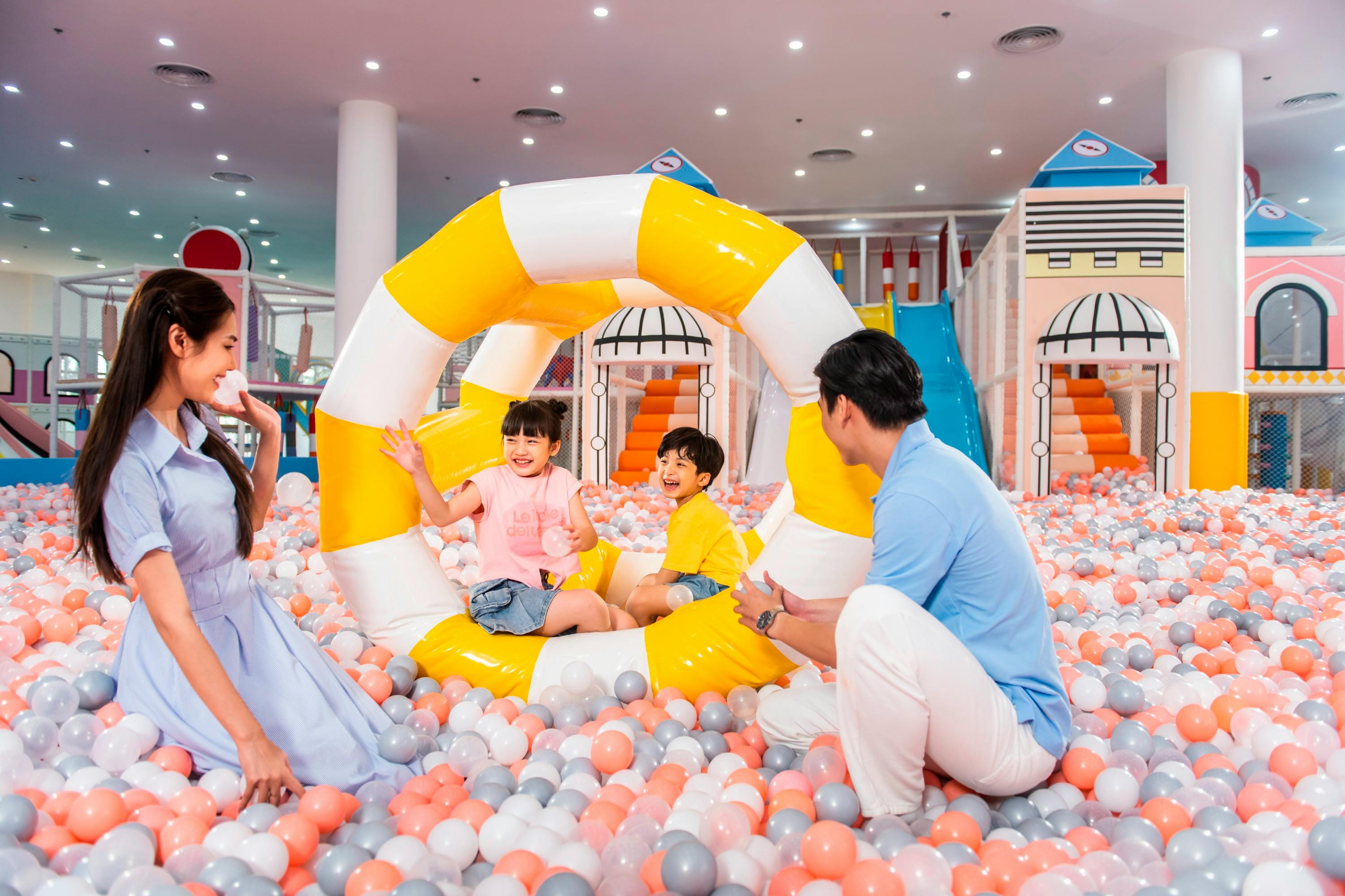 Children playing in a colorful ball pit with adults nearby, in an indoor play area with a yellow inflatable ring.