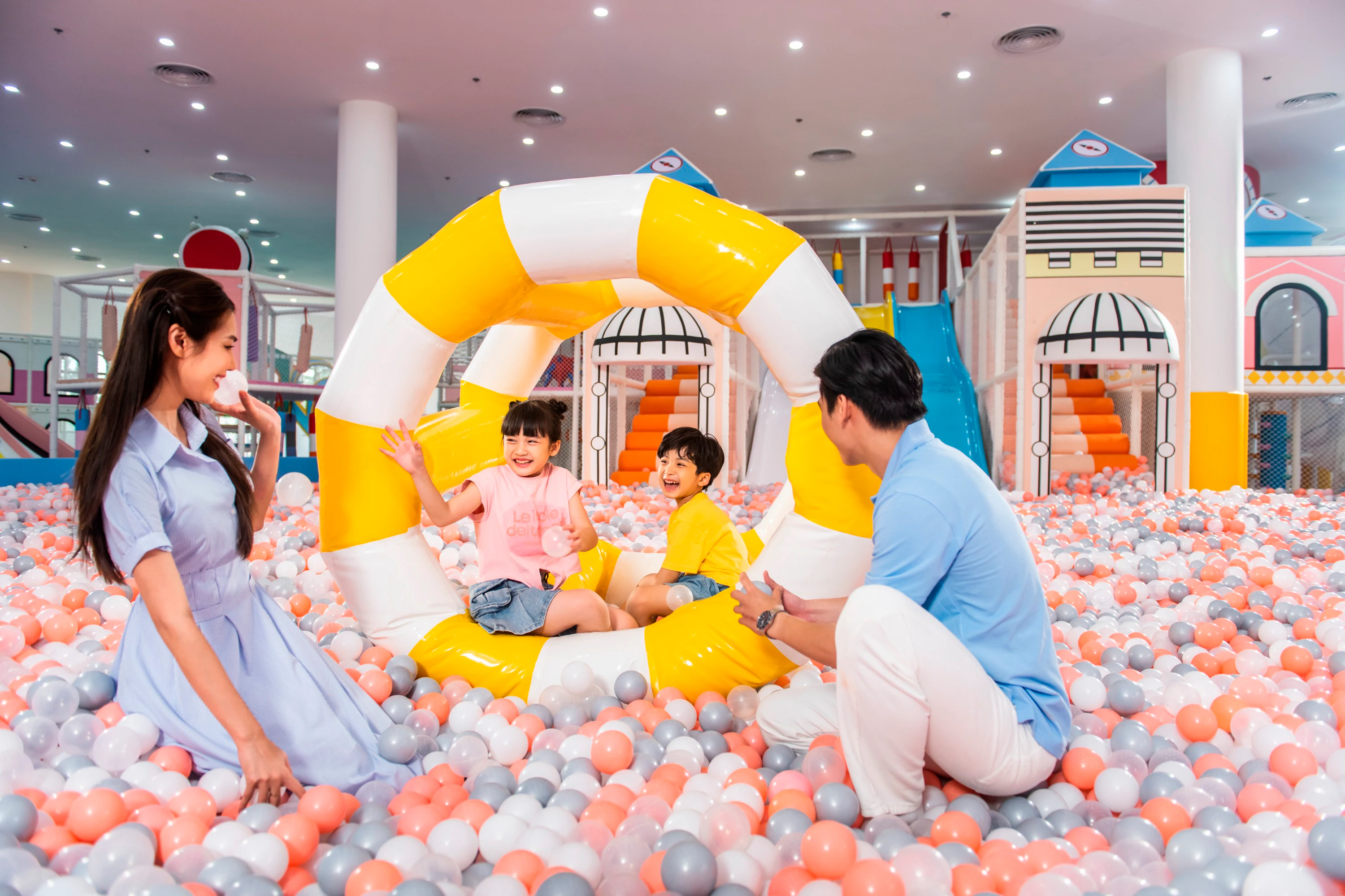 A family with two children playing in a colorful indoor ball pit with an inflatable yellow and white ring.