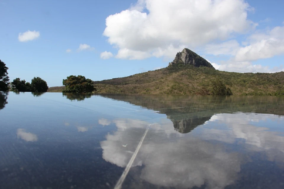 A reflective water surface mirroring a mountain and clouds under a bright sky, with greenery in the background.