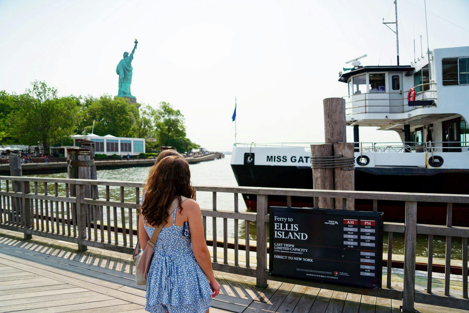 Een persoon in een blauwe jurk staat op een pier bij een veerboot die is aangemeerd bij een bord voor de Ellis Island veerboot. Het Vrijheidsbeeld is zichtbaar op de achtergrond.