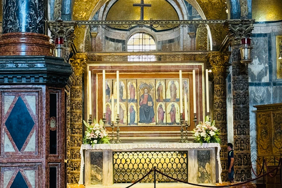 An ornate altar in a church with religious artwork, lit candles, floral arrangements, and a cross above.