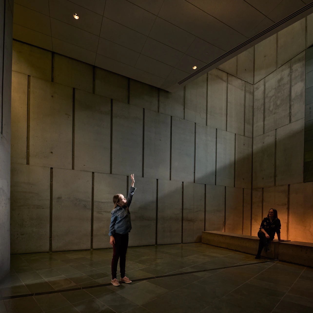 A person stands in a dimly lit, large room with high concrete walls, reaching up; another person sits on a bench along the wall.