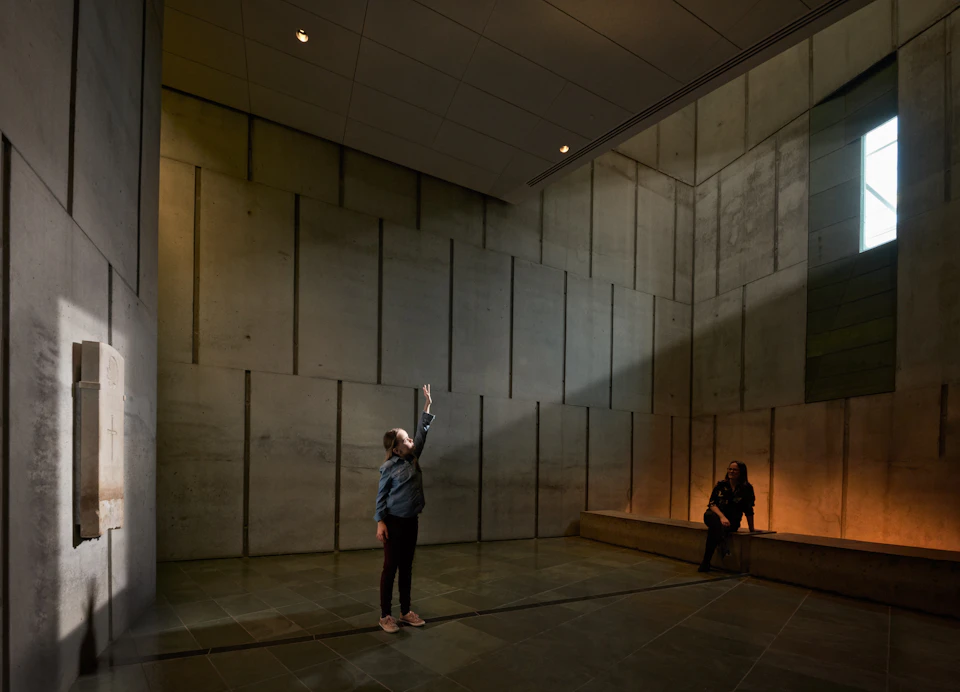 A person stands in a dimly lit, large room with high concrete walls, reaching up; another person sits on a bench along the wall.
