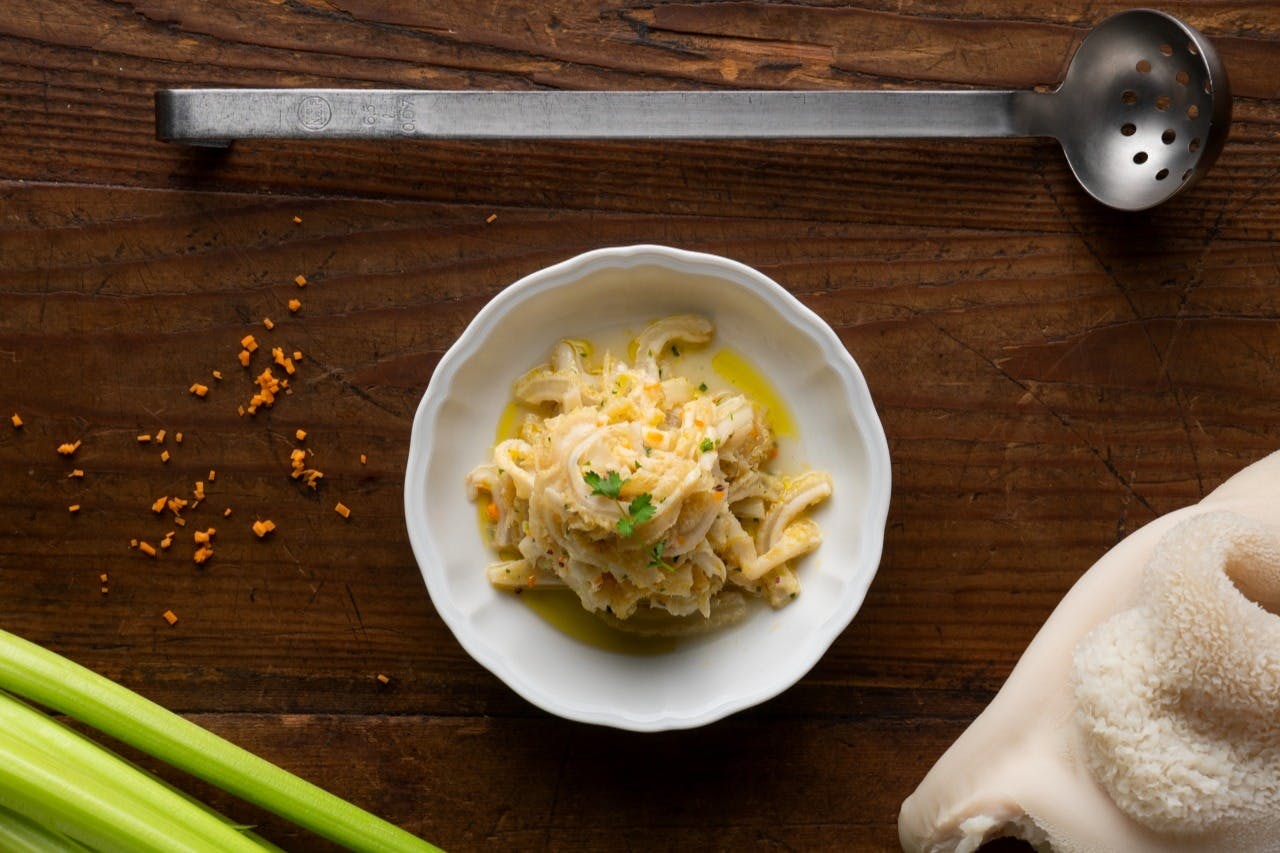 A white bowl of pasta with grated cheese and herbs on a wooden table, surrounded by cooking tools and ingredients.