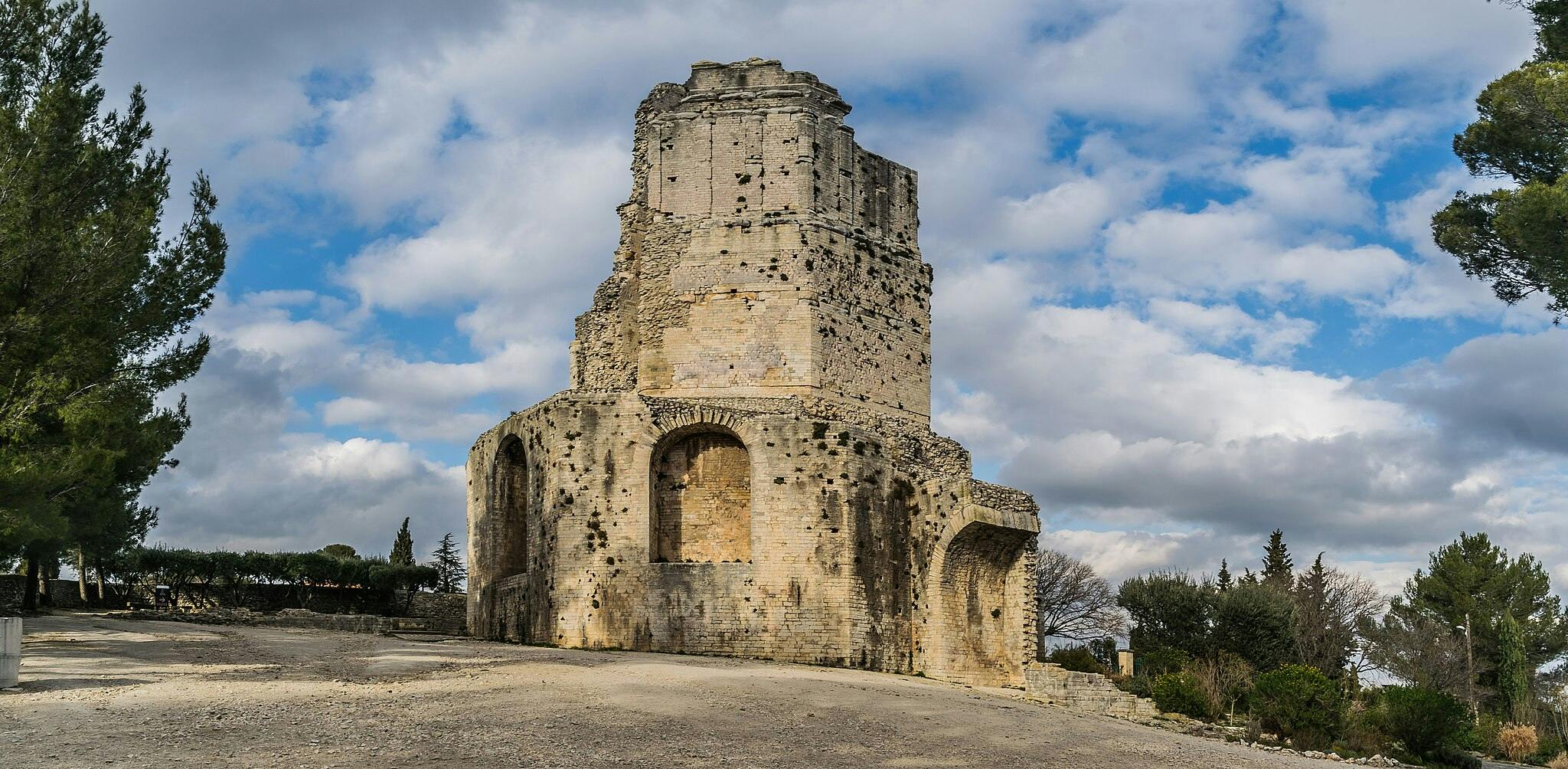 Ancient stone tower ruin under a partly cloudy sky, surrounded by trees and greenery, with a rocky foreground.