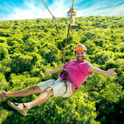 Ein Mann in einem rosa Hemd und einem Helm fährt mit einem Seilzug über einen dichten Wald und lächelt mit ausgestreckten Armen. Im Hintergrund sind ein paar Türme zu sehen.