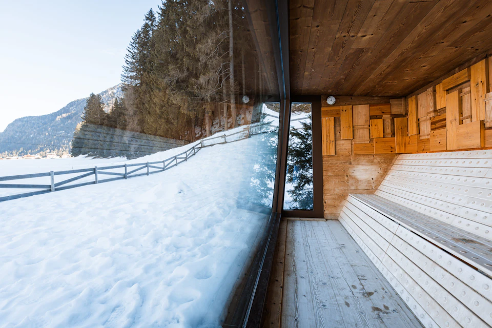 A wooden sauna with large glass windows overlooks a snowy landscape with trees and a wooden fence in the background.