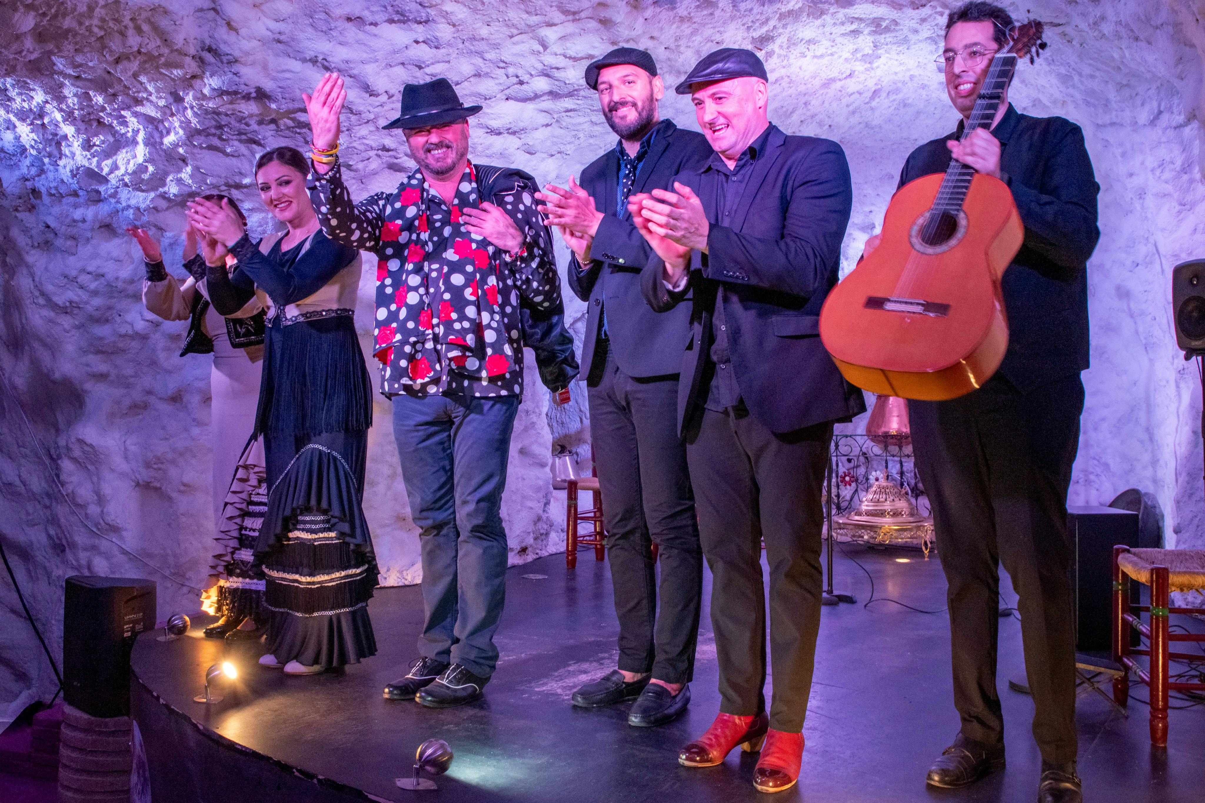 Four men standing on a stage, one holding a guitar, with a stone wall in the background.