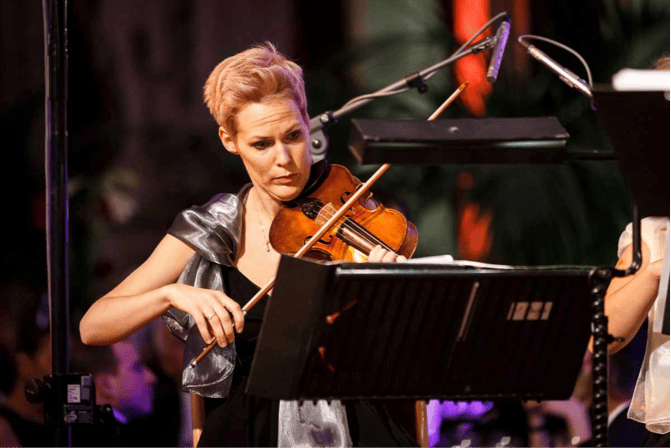 Ένα άτομο με κοντά μαλλιά που παίζει βιολί, φορώντας μαύρο φόρεμα και ασημί μαντήλι, επικεντρώθηκε σε ένα μουσικό περίπτερο κατά τη διάρκεια μιας παράστασης.