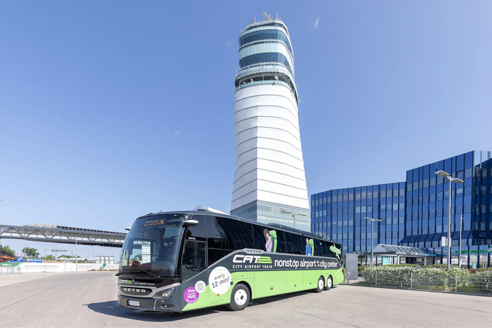 A green and black CAT bus labeled "nonstop airport-city center" is parked in front of a tall modern tower and glass buildings.