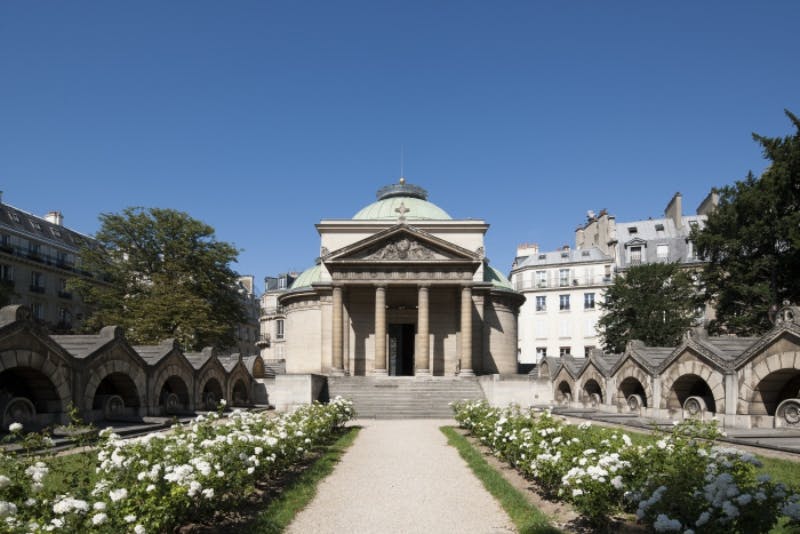 A classical building with columns and a dome, surrounded by white flowers and shrubs, set against a clear blue sky.