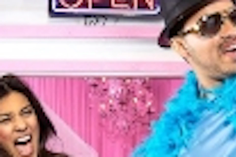 Two people enthusiastically pose in front of a colorful wedding chapel with neon signs, one wearing a blue feather boa.
