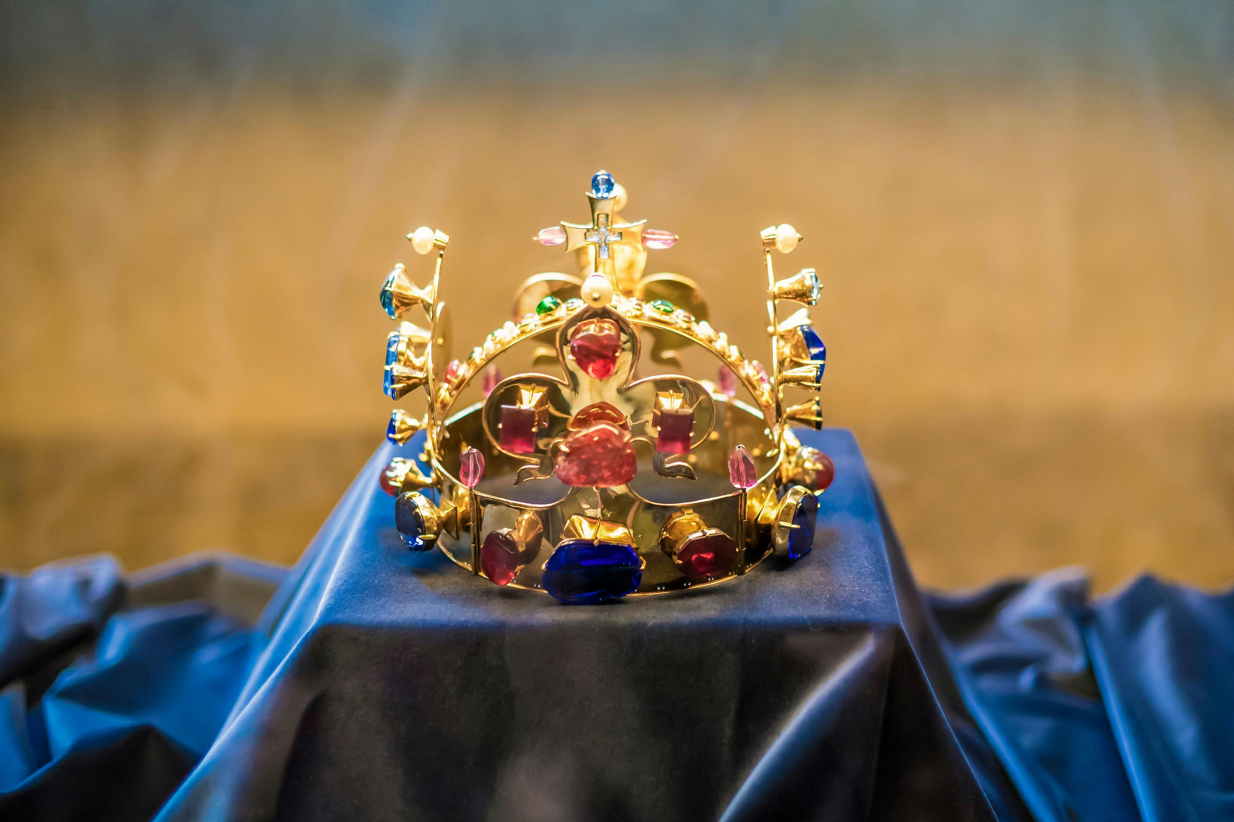 A gold crown adorned with colorful gems rests on a dark blue cloth display.