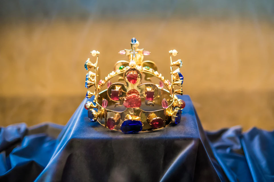 A gold crown adorned with colorful gems rests on a dark blue cloth display.