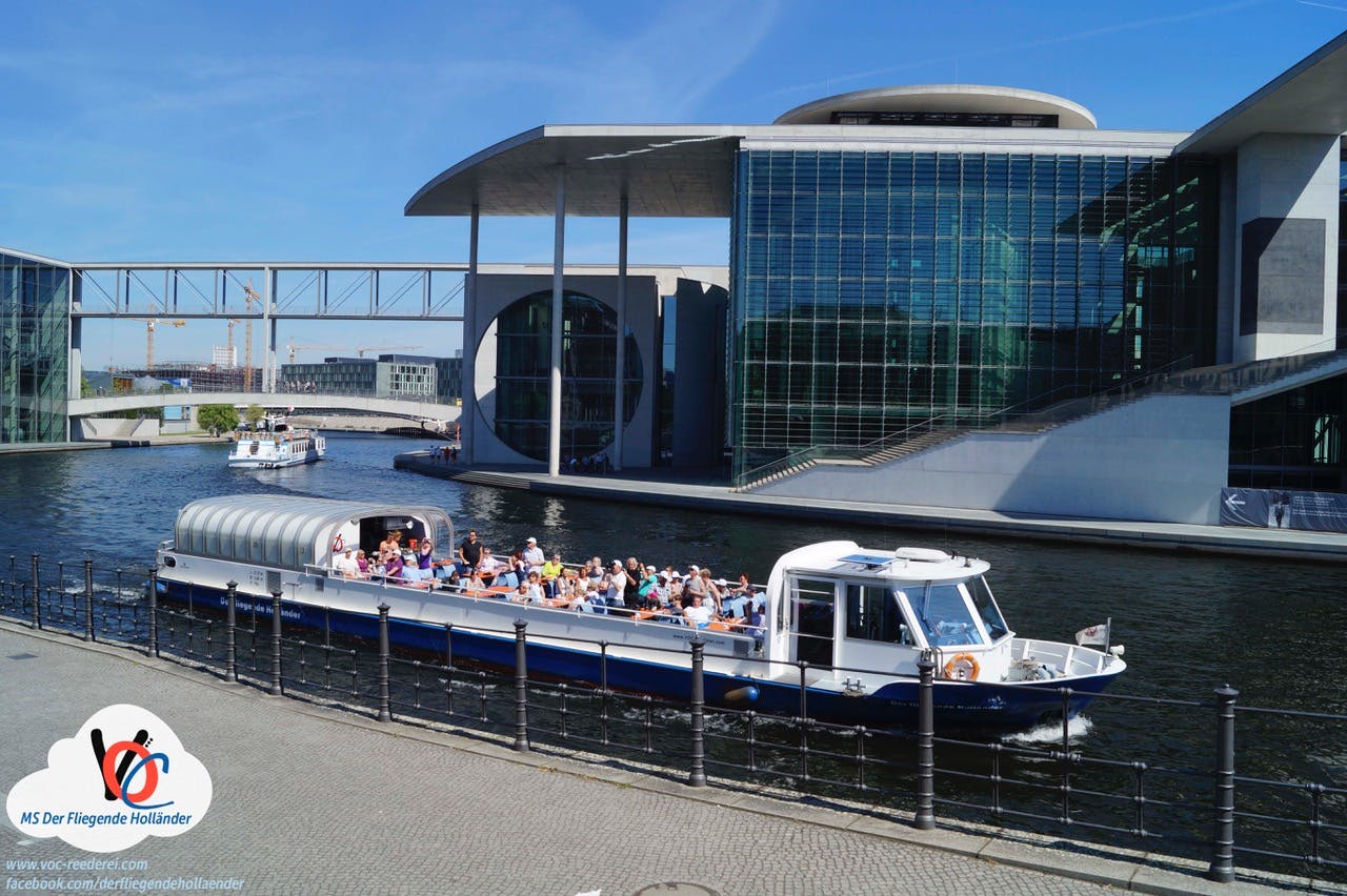 Ένα τουριστικό σκάφος γεμάτο ανθρώπους περνάει μπροστά από ένα σύγχρονο γυάλινο κτίριο και γεφυρώνει ένα ποτάμι μια ηλιόλουστη μέρα.