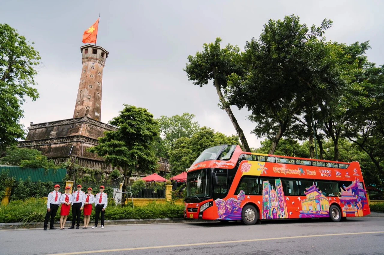 A red double-decker bus with colorful artwork is parked near a historic tower with a flag, surrounded by trees. Four people stand nearby.