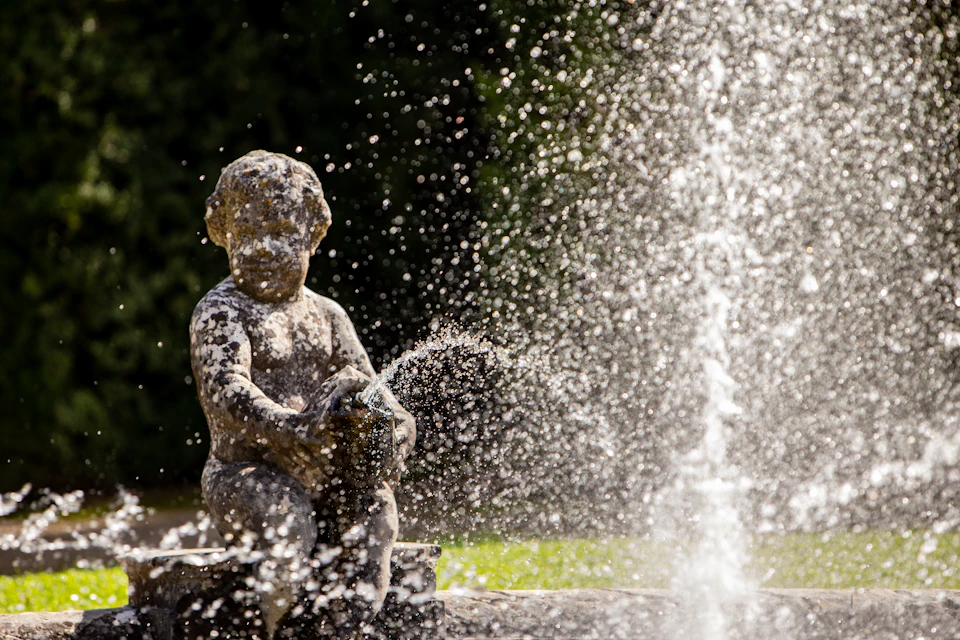 Stone cherub statue holding a spout, surrounded by water spray from a fountain, set against a dark green background.