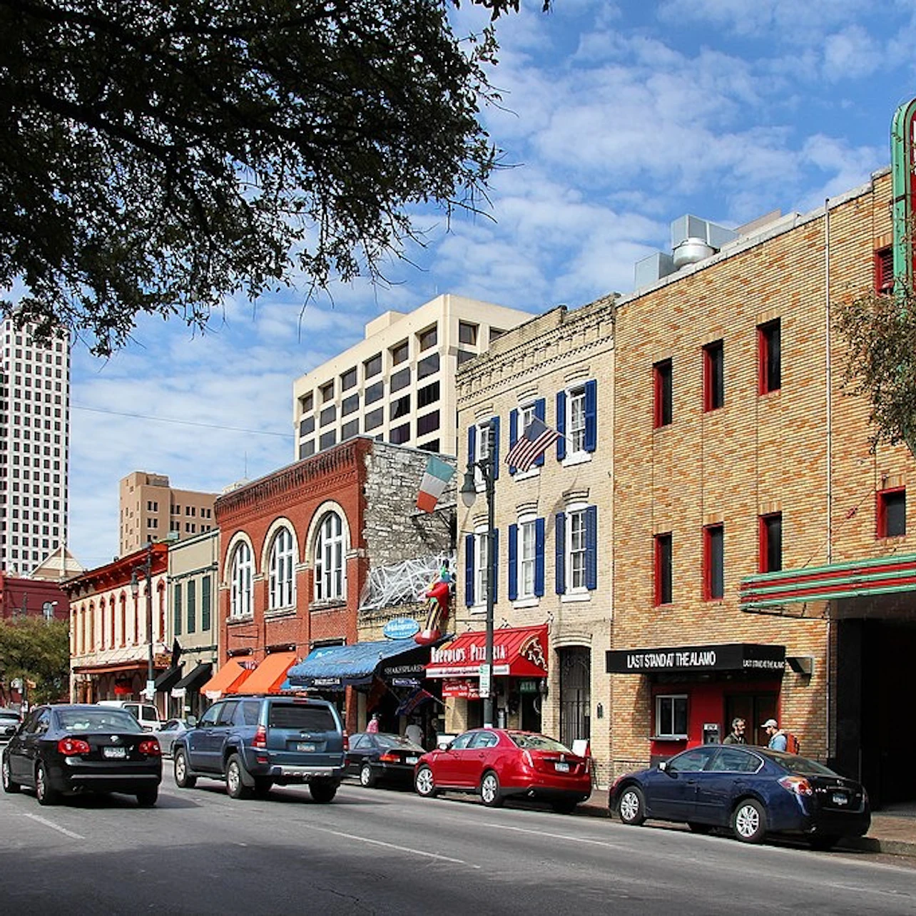A street scene with colorful buildings, parked cars, and pedestrians, with a clear blue sky and trees in the foreground.