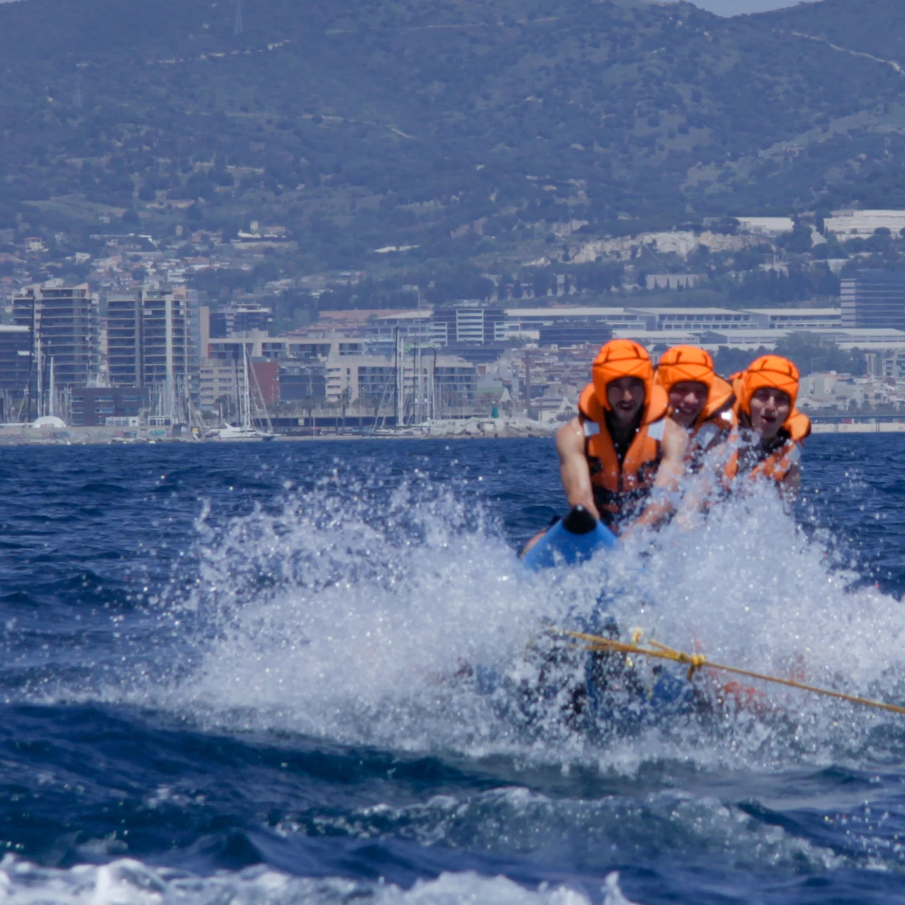 Four people in orange life vests ride a banana boat on the sea with a city and mountains in the background.