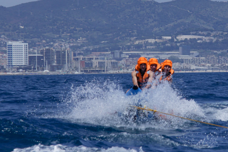Four people in orange life vests ride a banana boat on the sea with a city and mountains in the background.