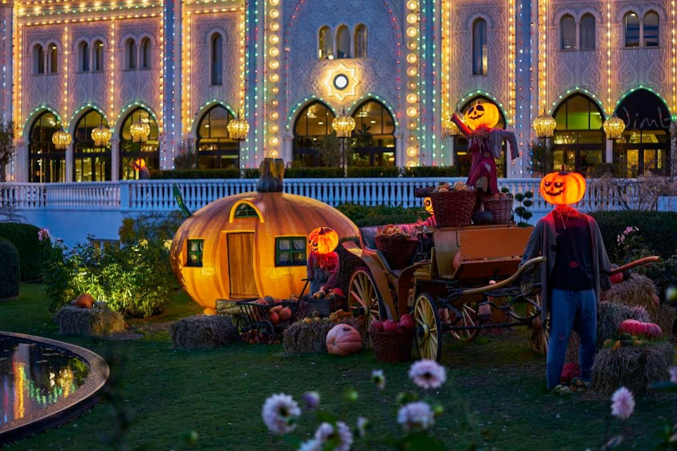 Halloween-themed scene with glowing jack-o'-lanterns, a pumpkin-shaped house, an old wagon, and a brightly lit building at night.
