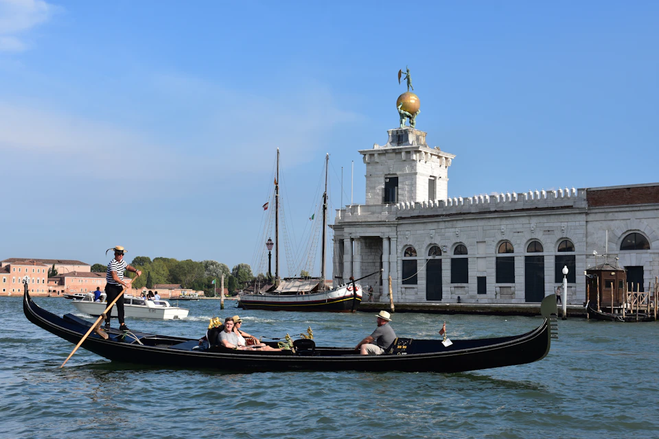 People riding a gondola on a canal with a historic building and other boats in the background under a clear blue sky.
