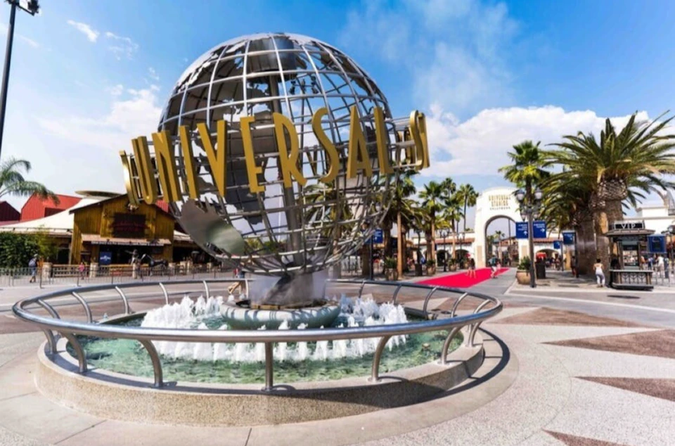 A metal globe sculpture with "Universal" text in a fountain at an outdoor area with palm trees and an archway in the background.