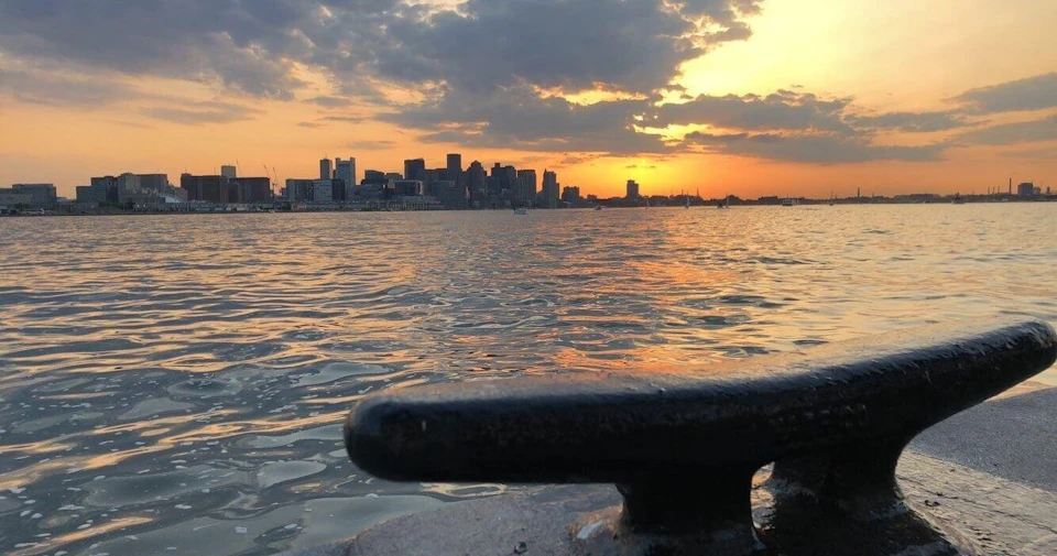 City skyline at sunset viewed across water, with a dock cleat in the foreground and clouds partially covering the sun.