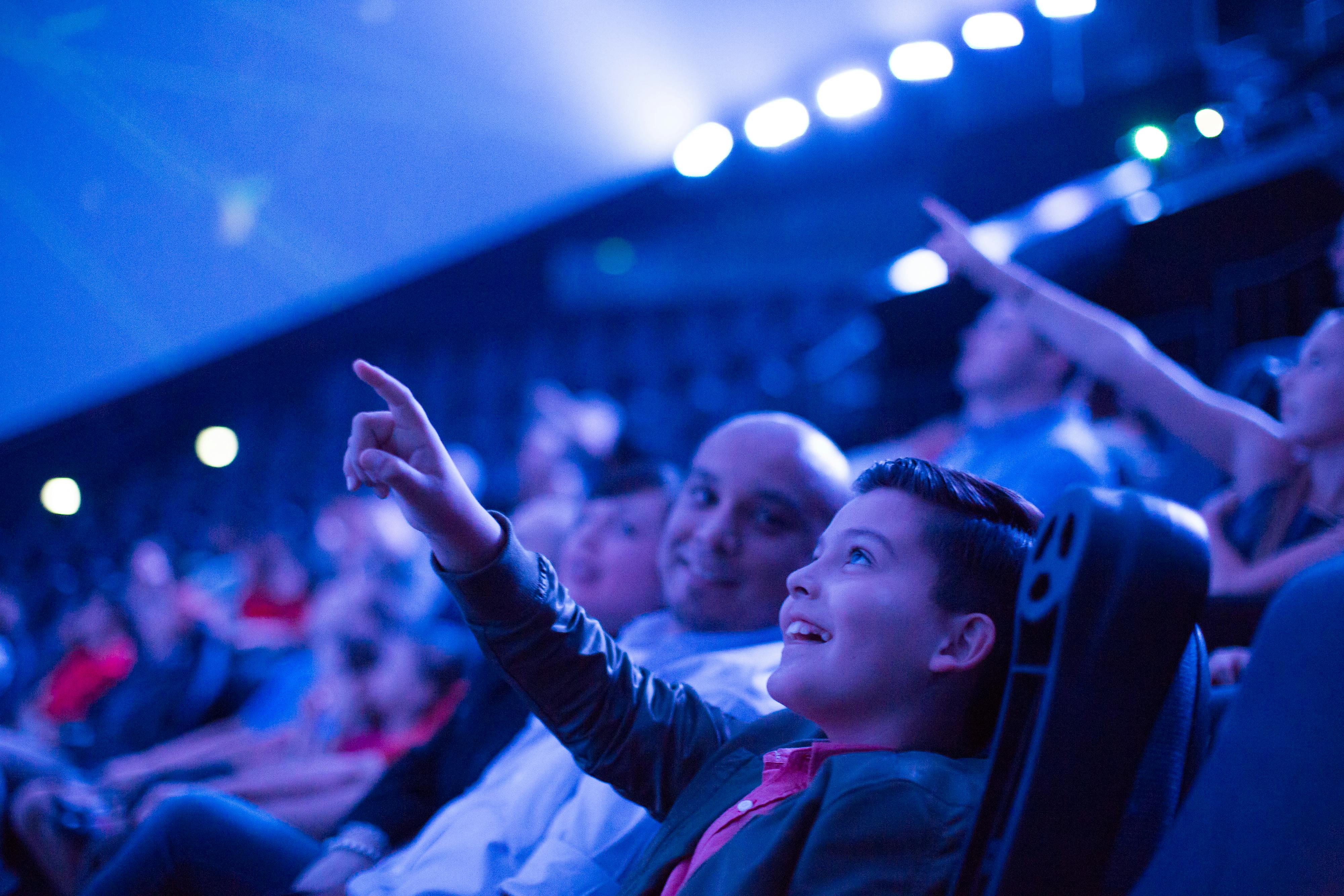 Άνθρωποι κάθονται σε ένα θέατρο, χαμογελώντας και δείχνοντας προς την οθόνη. Ο φωτισμός δημιουργεί μια μπλε ατμόσφαιρα.