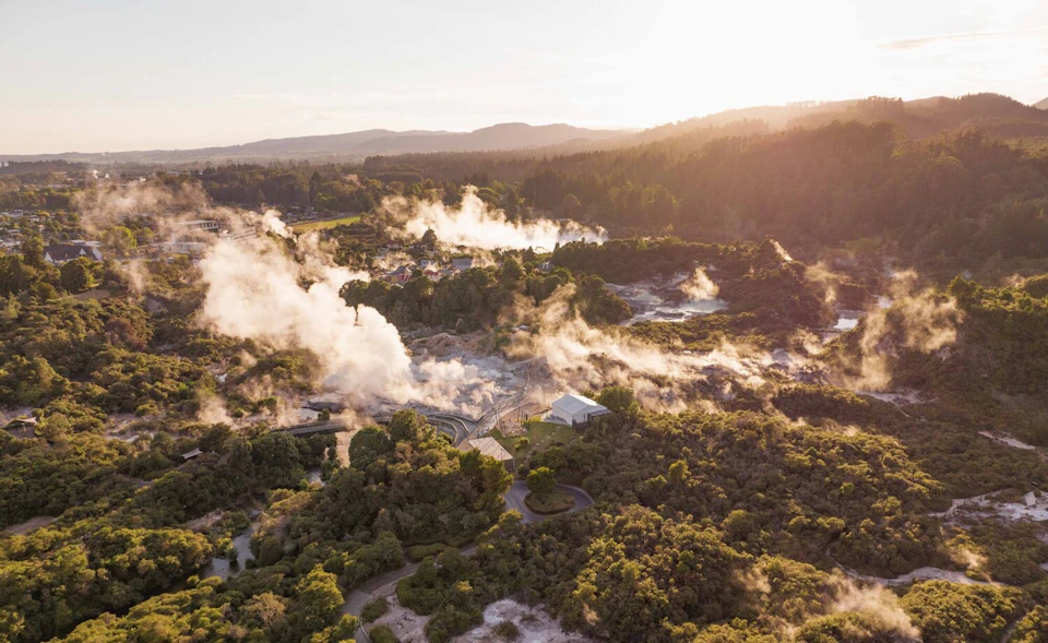 A geothermal area with steam rising from the ground, surrounded by vegetation and hills under a sunny sky.