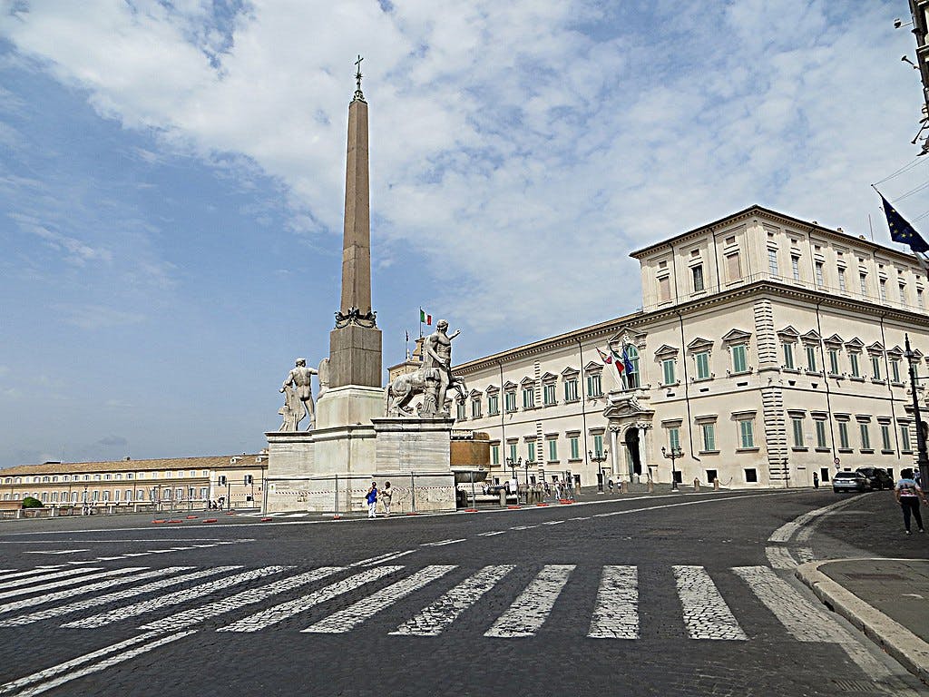 一座巨大的方尖碑矗立在鹅卵石广场上，周围环绕着部分多云的天空下的历史建筑。