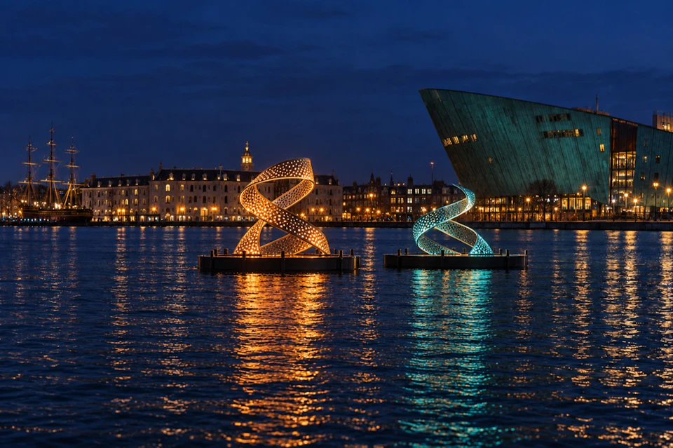 Two illuminated spiral sculptures, one orange and one blue, floating on water at night with buildings in the background.