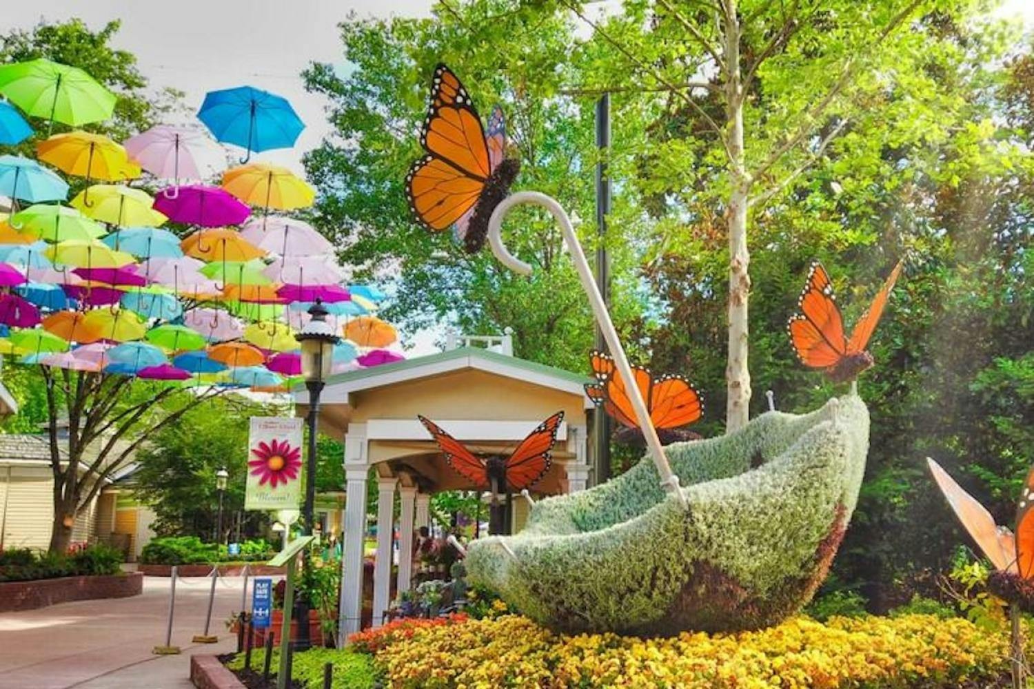 A whimsical outdoor scene featuring colorful umbrellas suspended in the air, a large butterfly sculpture, and lush greenery.