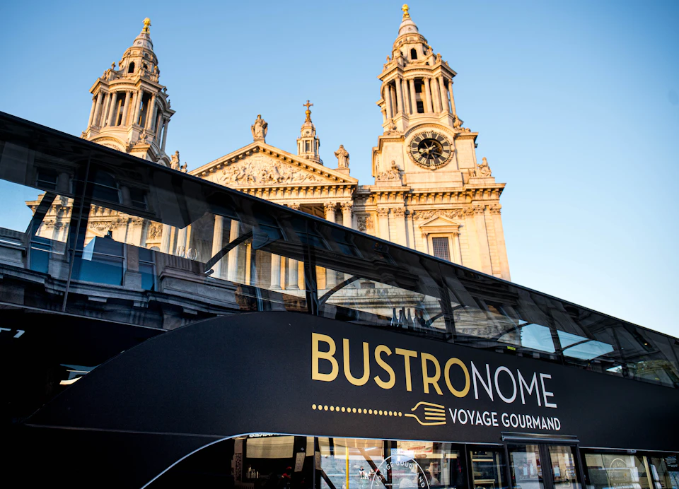 A double-decker tour bus labeled "BUSTRONOME VOYAGE GOURMAND" in front of a cathedral with twin towers and a clock.