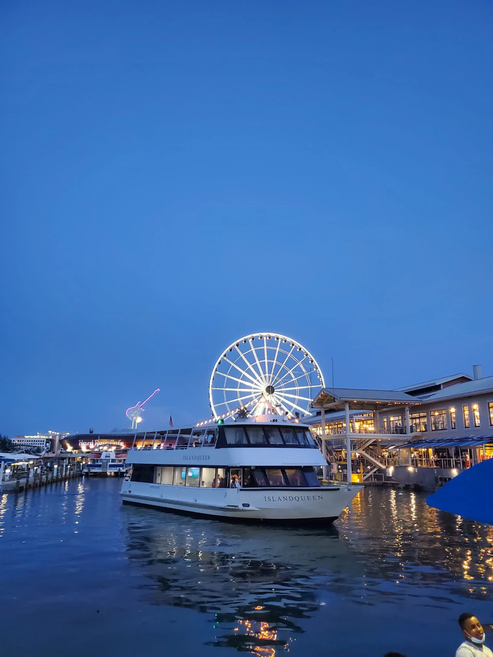 A boat labeled "ISLAND QUEEN" on a waterfront with a lit Ferris wheel and buildings in the background at dusk.